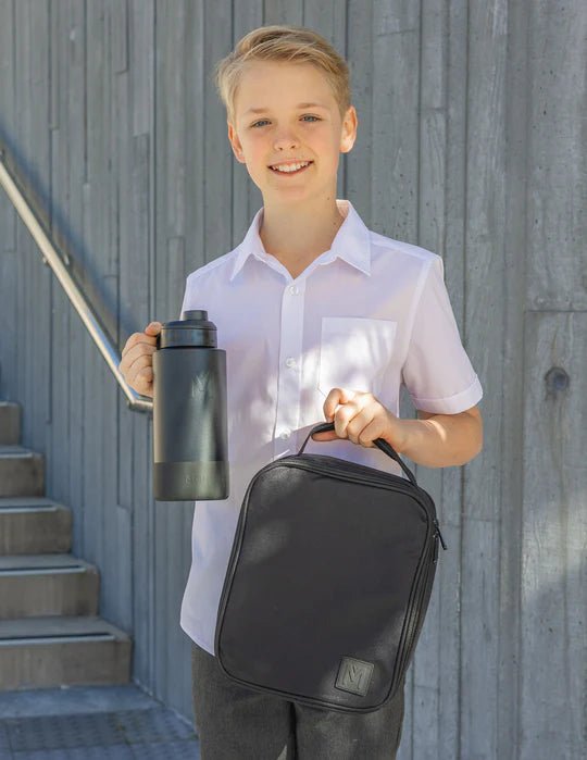 Young boy holding a black backpack and water bottle against a gray wall.